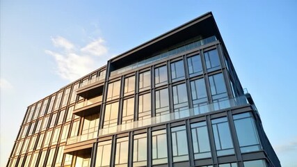 A low-angle shot captures a stunning modern glass building with a sleek, contemporary design, towering against a clear blue sky dotted with soft clouds. The reflective windows shimmer with dynamic con