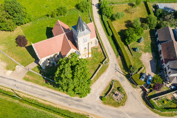 Drone shot of a historic stone church with a pointed steeple and red tiled roof, surrounded by green lawns, mature trees, and village roads in the rural setting of Cuncy les Varzy under bright