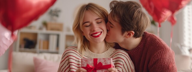 Young man kissing a woman on the cheek, holding a wrapped gift and heart balloons