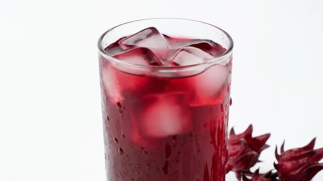 A vibrant, deep red or magenta-colored beverage, likely an herbal infusion or fruit drink, is presented in a tall glass filled with clear ice cubes. The glass is visibly frosted with condensation, ind