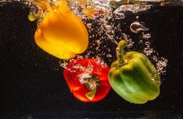 High speed underwater shot of yellow, green, and red bell peppers creating dynamic splashes against a dark background. This vibrant image represents freshness, healthy eating,