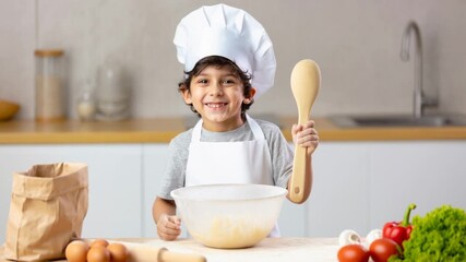 Child in Chef Hat Enjoys Baking With Mixing Bowl and Kitchen Ingredients as He Smiles and Poses With a Wooden Spoon