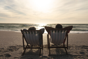 Retired senior multiracial couple relaxing while sitting on folding chairs looking sea from beach