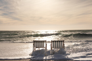 Wave splashing on empty two folding wooden chairs at beach against sky during sunset