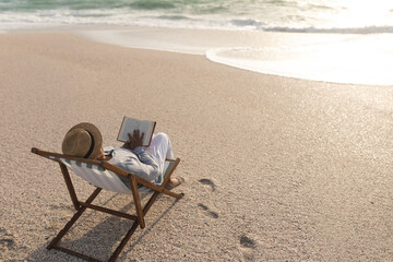 Relaxed senior woman reading book while sitting on folding chair at beach during sunset