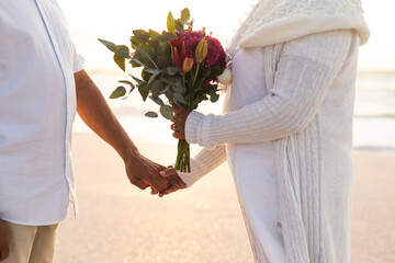 Midsection of multiracial senior couple holding hands with bouquet during wedding ceremony at beach