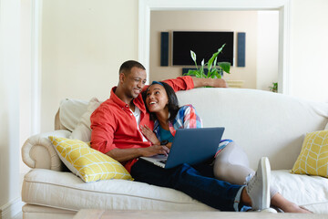 Smiling young african american couple lying on sofa with laptop in living room at home