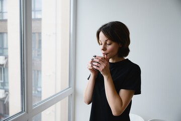 Young woman standing by a window and drinking a hot beverage in a bright minimalist interior. Morning routine, calm mood, comfort and slow lifestyle concept.