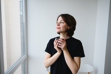 Young woman holding a cup of hot drink and looking out the window in a bright minimalist room. Calm morning mood, reflection, comfort and slow living concept.