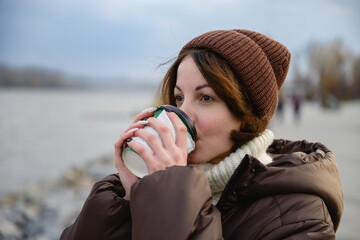Young woman wearing warm jacket and knit hat drinking hot coffee from a takeaway cup outdoors on a cool autumn day. Seasonal lifestyle, comfort, relaxation and urban nature concept.