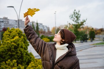 Young woman wearing warm clothes holding a yellow autumn leaf in an urban park. Fall season, calm mood, outdoor lifestyle and connection with nature concept.