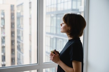 Thoughtful woman holding a cup of coffee while looking out a window at an urban cityscape. Quiet morning moment, mindfulness, lifestyle and calm reflection at home.