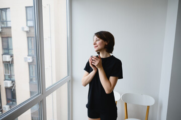 Thoughtful woman holding a cup of coffee while looking out a window at an urban cityscape. Quiet morning moment, mindfulness, lifestyle and calm reflection at home.
