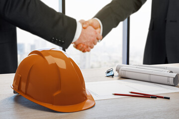 Businesspeople shaking hands over construction deal with hard hat and blueprints on office desk in modern high-rise background setting.
