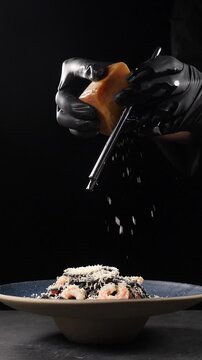 Italian French cuisine, cheese being grated on black pasta shot on black background, restaurant food cooking and serving. Vertical footage. Chef in black gloves grating delicious hard cheese on grater