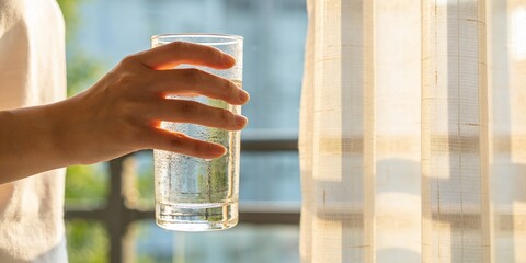 Close up of woman hand holding fresh glass of pure water with condensation near window with sunlight, healthy lifestyle and hydration concept for wellness and skin care