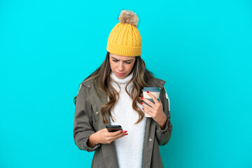 Young Italian woman wearing winter jacket and hat isolated on blue background holding coffee to take away and a mobile