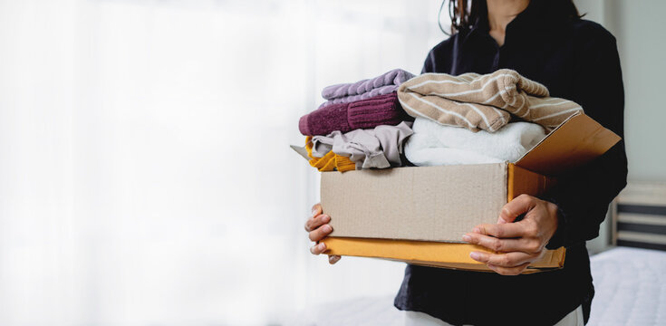 Female hands holding a cardboard box filled with used clothing. Concept of clothes donation, charity, sustainability, recycling, and home decluttering.