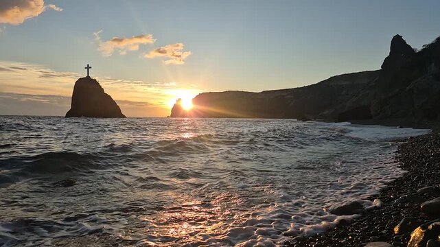 Cliff sunset ocean waves crashing on a pebble beach near the large holy rock with a cross. Vertical video