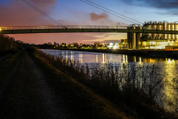 The Gersthofen industrial park, a chemical and industrial site with futuristic-looking buildings and silo facilities. The green bridge construction spans the Lech Canal. Colorful cloudy evening sky