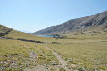 A long narrow walking path stretches across a vast grassy field between the rocky hills of Mala Luka and Vela Luka on Krk island