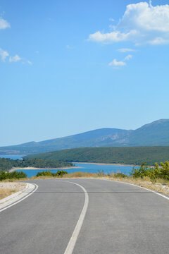 A quiet asphalt road curves through the scenic Dalmatian hinterland on the eastern side of Peruca Lake with mountains and water visible under a blue sky