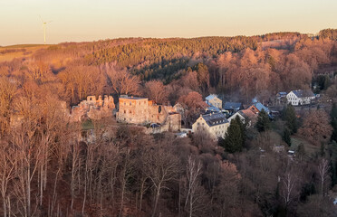 Aerial view of a historic castle ruin in the city of Hartenstein, saxony, nestled among autumn-colored trees, surrounded by a small village and bathed in warm evening sunlight