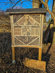 handcrafted insect hotel stands in a leaf-covered field, illuminated by bright winter sun, offering shelter for beneficial insects amid bare trees and dry grass