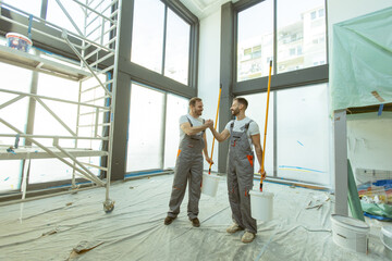 Workers shake hands while painting a room in a large, sunny space with scaffolding
