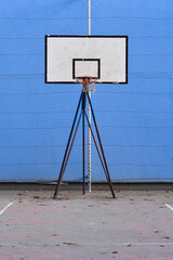 A full front view of a white basketball backboard and hoop on a metal stand located on an outdoor concrete court with a blue safety net background