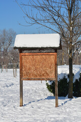 A wooden notice board with a snowy roof stands in a public park surrounded by deep white snow under a clear blue sky