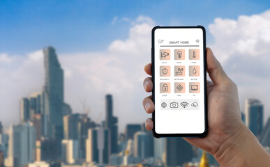 Hand holding smartphone with smart home control app against blurred urban city skyline. IoT technology and home automation concept.