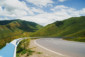 Winding asphalt road through lush green mountains under a bright blue sky with fluffy white clouds