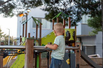 Obraz premium Back view of blonde boy in striped shirt standing on playground platform looking at colorful play structures in resort park. Childhood curiosity, outdoor exploration, summer fun and leisure.