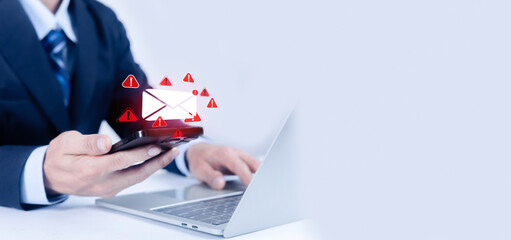 A businessman uses a laptop and smartphone with a digital envelope and red warning icons floating above. This image depicts cybersecurity threats, phishing, and urgent alerts in a corporate setting.