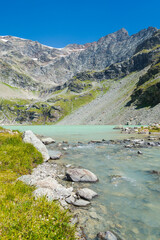 Verfical view of the "San Grato" lake, Aosta Valley, Italy, with water flowing in the foreground. High peaks on the background with blue sky.
