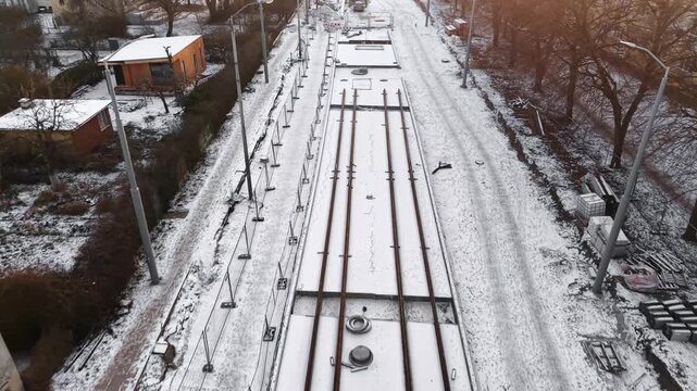 Aerial drone view of snow-covered tram tracks under construction &mdash; slab-track rails on concrete with fencing, utilities and workers along a winter urban corridor (forward flyover)