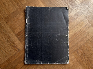 Vintage magazine mockup without cover, featuring heavy wear, cracked edges, and visible print lines. The blank black magazine lies flat on a wooden parquet floor.