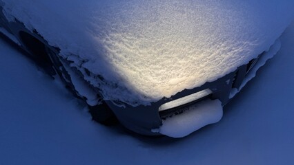 High angle view of a car hood and bumper buried in snow with headlights turned on. Warm light illuminating the snowy texture during a blue winter evening