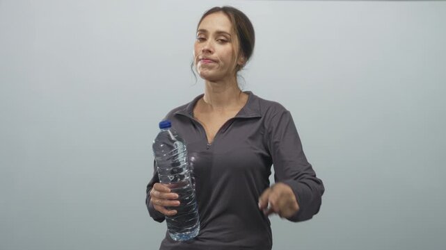Woman points finger while holding large water bottle, wearing dark gray zip top and looking at camera in studio; hydration reminder assertive.