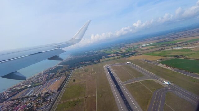 airplane wing sweeps active runway complex taxiways during departure patchwork farmland coastal water spread out under hazy blue sky aerial view airport flight 