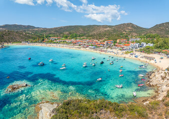 Aerial view of Kalamitsi Beach in Sithonia, Greece, with turquoise waters, golden sand, beach umbrellas, rocky islets, and a traditional seaside village.