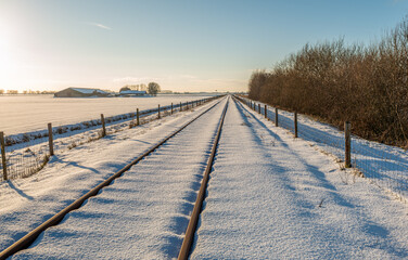 Snowy polder landscape with train tracks and a farm at the end of a sunny winter day. The photo was taken in the Dutch province of North Brabant.