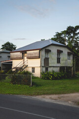 A side-angle view of a modest, elevated house in a lush tropical setting, featuring weathered siding and a classic silver corrugated metal roof. 