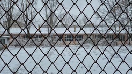 A snow-covered yard and a long, low building with many windows are seen through the diamond patterns of a rusty chain-link fence on a gray, overcast winter day.