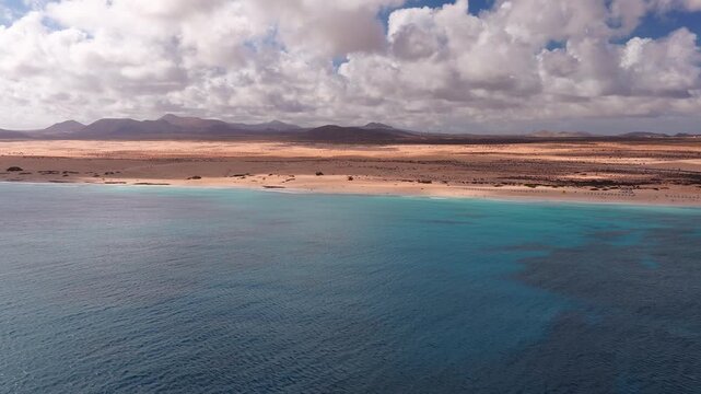 Aerial view of Fuerteventura, Canary Islands, shows white sand beach, golden dunes, gentle waves, umbrellas, wind turbines, volcanic peaks, and patchy sunlight.