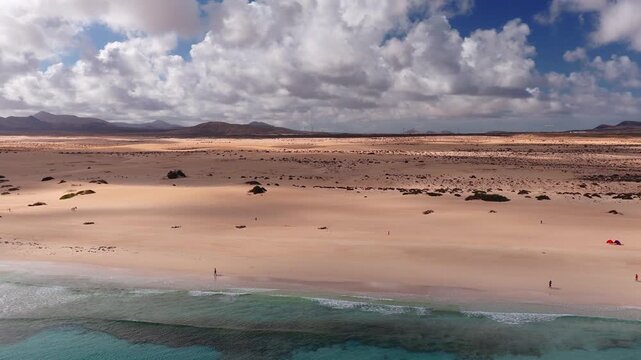 Aerial view of Fuerteventura in the Canary Islands, dunes and scrub, tiny people, a red tent, turquoise Atlantic, reefs, low volcanic hills, and wind turbines.