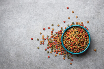 A blue bowl filled colorful dry pet food kibble with scattered on gray stone textured background. © PixelArt_25