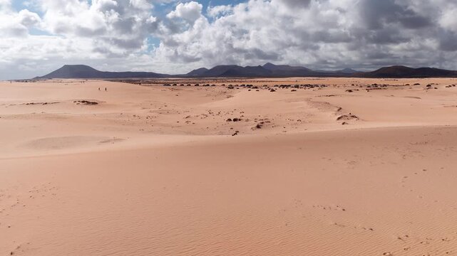 Aerial view of Corralejo dunes in Fuerteventura under low clouds. Rippled sand, scrub, footprints, parked cars, tiny walkers, and a flat topped cone are visible.