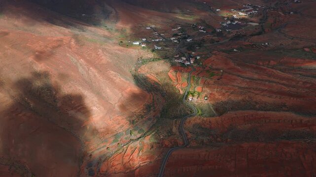 Aerial view of Fuerteventura, Canary Islands, with a winding road, whitewashed houses, layered terraces, irrigated plots, cloud shadows, and roadside vehicles